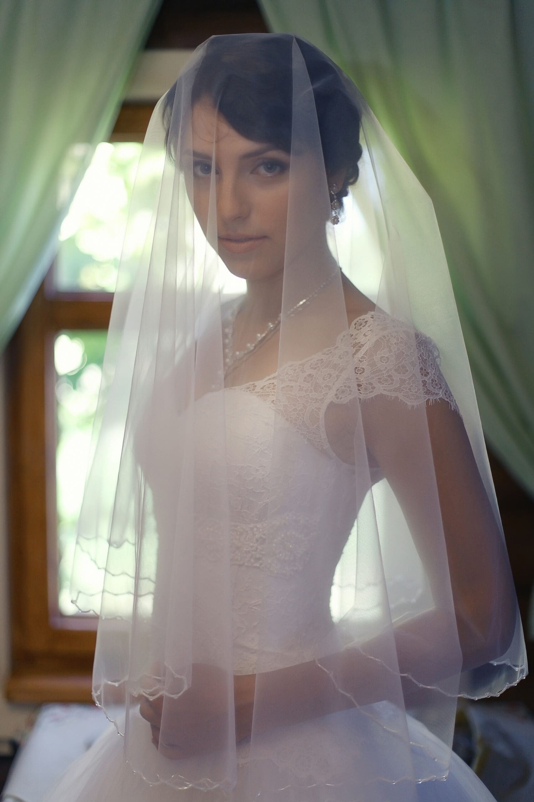 Bride standing indoors wearing a short, sheer wedding veil over her face